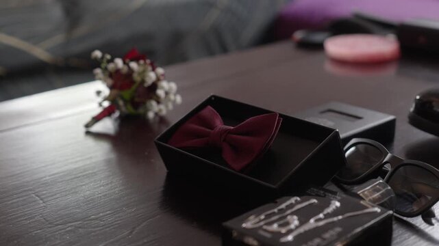 Red bow tie in a presentation box with boutonniere and glasses arranged on a table, flatlay styling as hands grab piece