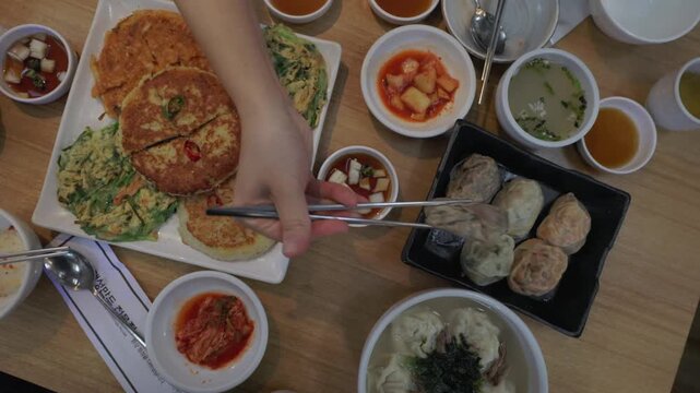 Table Filled With Korean Dishes. Savory Pancakes (Jeon) And Assorted Dumplings (Mandu) Being Picked Up With Chopsticks. High-angle Shot