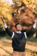 Fototapeta premium Cheerful man with arms raised amid swirling autumn leaves for fall banner and seasonal background featuring warm golden light and nature
