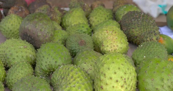 Green soursop fruit fills a detailed close view showing its uneven spiked skin while more tropical produce and the edge of a market display remain softly visible behind in daylight