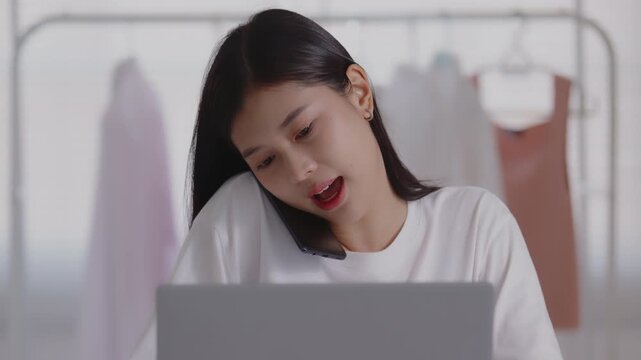 Young Asian woman talking on a smartphone while working on a laptop at her desk in a clothing studio, managing online business and customer service.