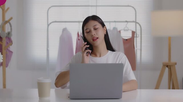 Young Asian woman talking on a smartphone while working on a laptop at her desk in a clothing studio, managing online business and customer service.