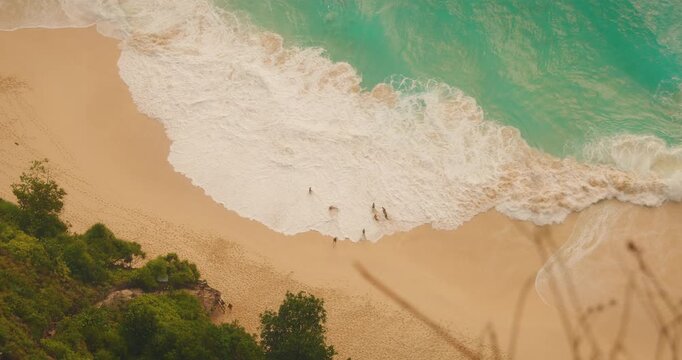 Top down view of blue ocean waves and people relaxing on white sand at Kelingking beach Bali Nusa Penida