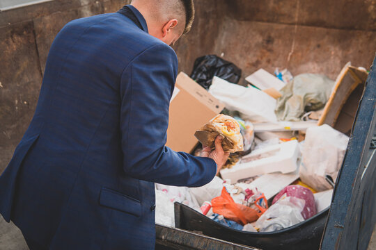 an impoverished businessman rummages in a trash can in search of food. A homeless homeless man in a suit holds burger in his hand while standing in a landfill. bankruptcy, no money concept