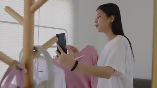 Young Asian woman holding a smartphone and showing a white shirt on a hanger during a live stream, presenting products for online business and e-commerce sales.