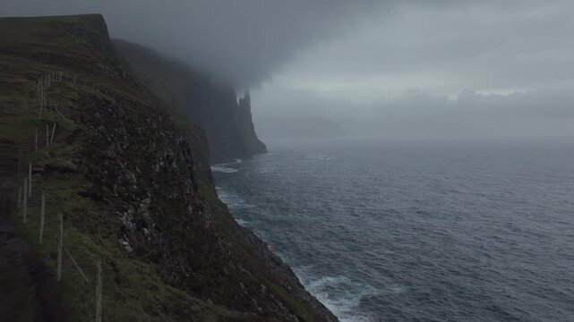 Dramatic aerial footage revealing the Trollkonufingur sea stack on a foggy day. A lone hiker admires the moody North Atlantic scenery