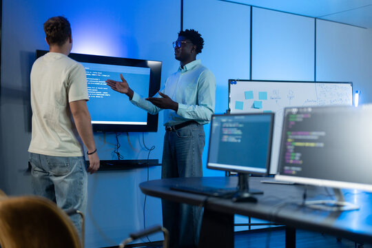 African American IT manager giving technical briefing to Caucasian developer in modern offices. Diverse male programmers discussing software presentation on screen. Corporate technology teamwork