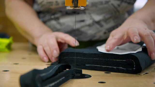 A close-up of fabric being cut. A utility knife carefully cuts a stack of fabric. The seamstress's hands hold the fabric firmly on the wooden cutting table, ensuring a straight and precise cut.