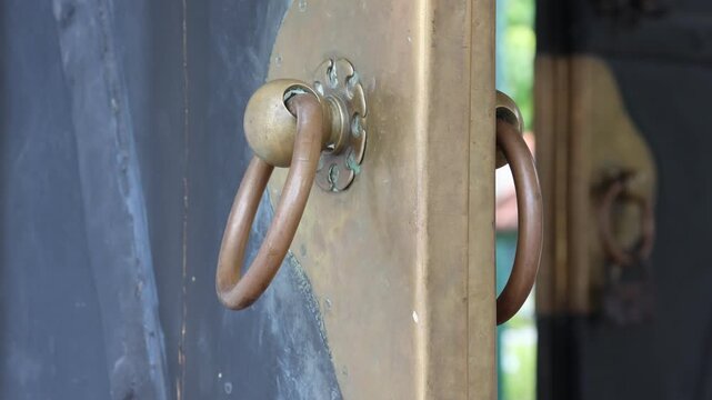 Old brass door with antique ring and padlock. Weathered metal surfaces show time and use. Dark door contrasts with golden hardware. Lock hangs securely on vintage door handle