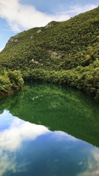 Vertical aerial drone view of the green water reflecting a forested mountain and the clouds above it, Soca Valley, Slovenia