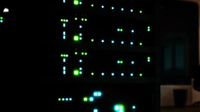 Several server units with active lights are visible in a dark room. The glow from the lights shows the status of the servers. An office chair and a plant are also present in the background.