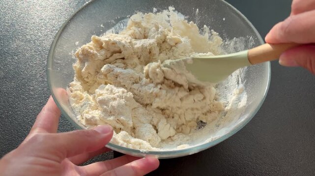 Female hands kneading raw dough in a glass bowl on a dark countertop. The hands-on process of baking