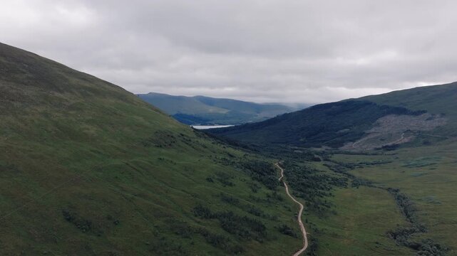 Aerial view of a narrow footpath winding up a green hillside toward a distant loch.