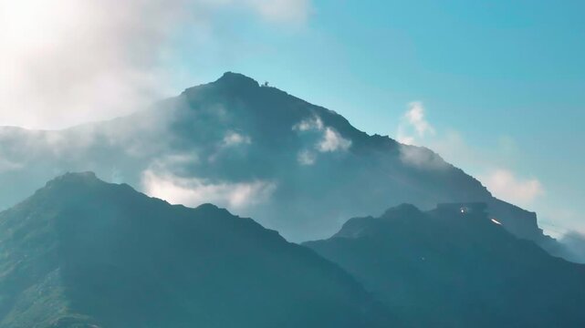 Aerial shot of a mountain summit rising above a dramatic sea of clouds, with mist wrapping around the peak under a clear blue sky.