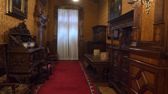 Pan shot of an ornate hallway inside Peles Castle featuring dark carved wood cabinets golden damask wallpaper and red carpet in Sinaia.