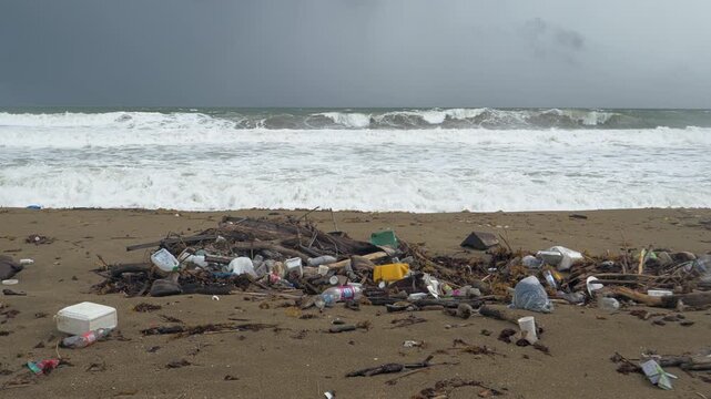 Mixed plastic containers bottles foam packaging bags and driftwood form a concentrated pile on open sand while broad white storm waves break across the entire ocean background