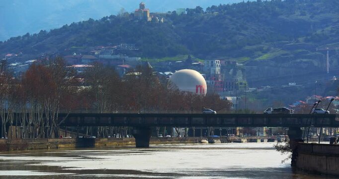 Traffic crosses a bridge over the Mtkvari River in Tbilisi while a white balloon and riverside buildings sit below a historic church on a green hillside