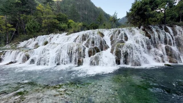 Jiuzhaigou slow motion shot of clear turquoise water cascading over mossy calcified rocks at Double Dragon Lake Waterfall in Sichuan, China, with dynamic splashes and forest scenery. Shuang Long Hai