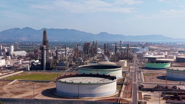 Large oil refinery complex with massive storage tanks, distillation towers and industrial infrastructure stretching across a coastal plain with mountains in the background