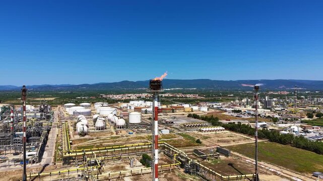 Large oil refinery complex with flare stack burning gas, storage tanks, pipelines and industrial facilities spread across a wide area with mountains and blue sky