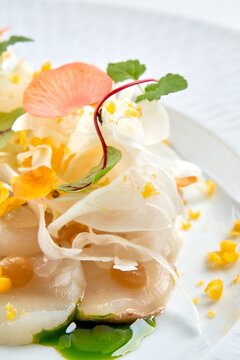 Macro shot of scallop carpaccio with green herb oil, shaved fennel, and edible flower petals on a white plate.