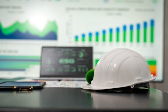 White safety hard hat on a desk beside a tablet and pen, with blurred green business charts on a screen in the background.