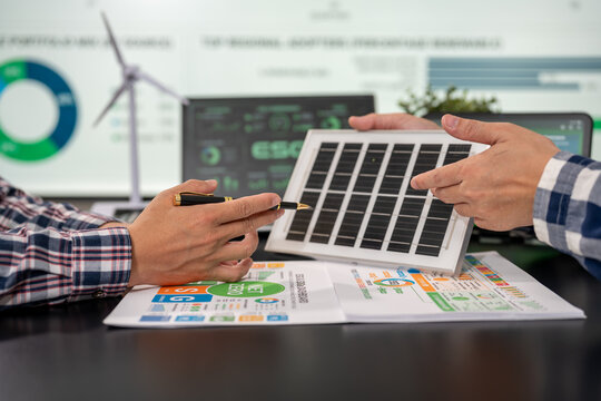 Two people discuss a small solar panel over renewable energy charts and a laptop, with a wind turbine graphic in the background.