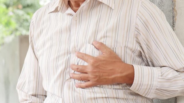 elderly asian man with silver hair suffering from cardiac pain outdoors, grimacing and holding chest in a park