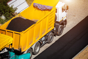 Aerial view of heavy construction machinery, large yellow dump truck delivering fresh asphalt for road paving, new road surface formation in progress under bright sunlight. Copy space © HENADZY