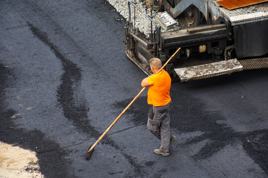 Man worker rakes hot asphalt surface for road construction project. Heavy machinery in background prepares for laying fresh pavement. Road building process for infrastructure development. Copy space