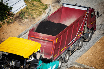 Heavy-duty red dump truck transporting and unloading hot asphalt into road paver machine at construction site for road building project. Aerial view of paving process. Copy space © HENADZY