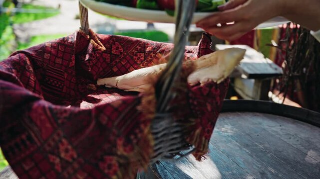 Hands lift a plate of fresh vegetables and herbs from a wicker basket lined with a red carpet and set it on an old wine barrel.