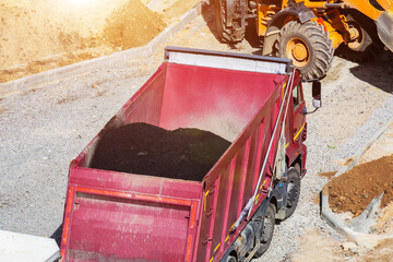 Red dump truck hauling asphalt material on road construction site, heavy machinery and earth piles visible, work in progress of new infrastructure paving project development. Copy space © HENADZY
