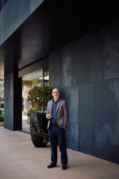 Mature businessman using cell phone while standing in front of office building and looking at camera.