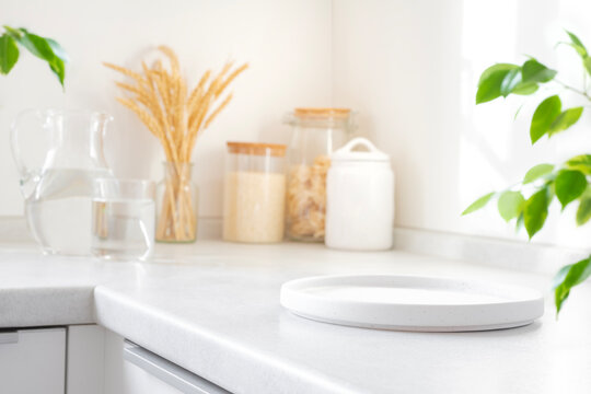 Empty white plate close-up on kitchen counter with blurred glass pitcher, jars of grains and green leaves as background