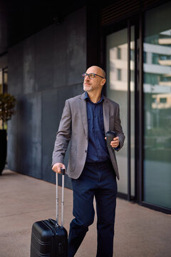 Mature businessman walking in city with takeaway coffee and suitcase.