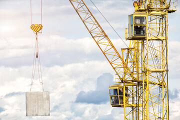 Yellow construction crane lifting heavy concrete panel against cloudy sky at building site, showing intricate metal structure of boom and operator cabin © HENADZY