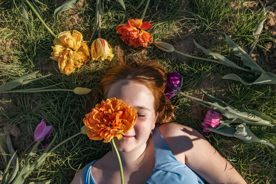 A girl lies on grass holding an orange tulip over her face