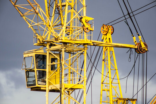Close-up view of bright yellow metal structure of a large construction crane and operator cabin, with pulleys and cables prominent against a gray, cloudy sky, building site equipment.