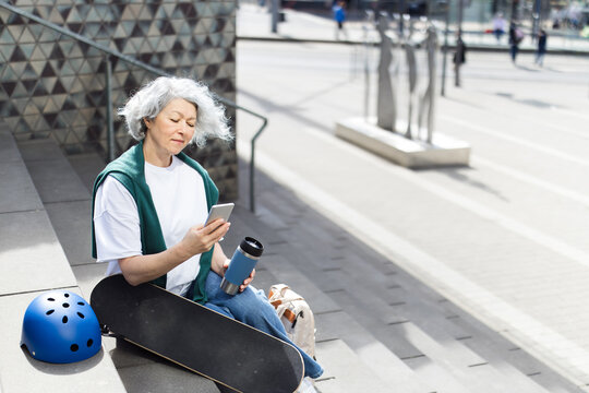 Woman sits on city steps using a smartphone next to her skateboard