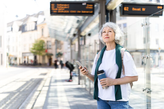 Woman waits at city tram stop holding smartphone and coffee cup