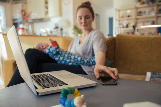 Mother with newborn baby at home taking cell phone from table