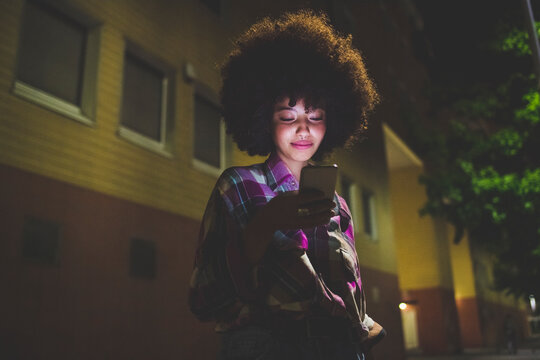 Young woman with afro hairdo using smartphone in the city at night