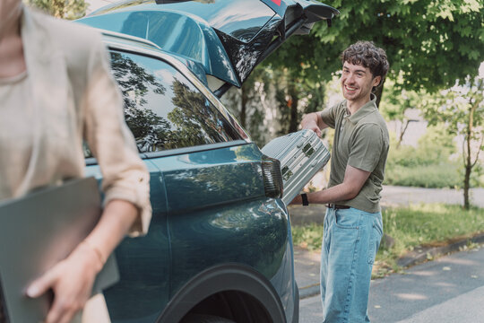 Smiling young man loading suitcase into car trunk while woman walks by
