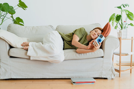 Tired woman relaxes on sofa and uses smartphone at home
