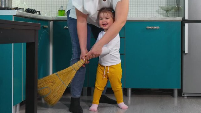 Little girl sweeps the kindergarten floor with help from a young Caucasian educator. The practical learning activity promotes independence, motor skills, confidence, and early childhood development