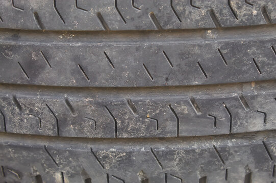 Close up of a used car tire showing detailed tread patterns, worn rubber surface, dirt, and textured grooves, highlighting the condition and structure of an automotive tire.