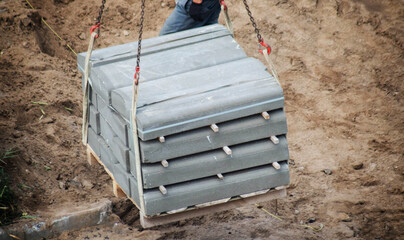 Fototapeta premium Concrete curb stones on pallet lifted by manipulator chains for construction work. Road parts transported over brown earth. Infrastructure development material movement unloading process.