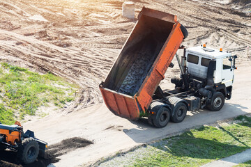 Dump truck tipping asphalt material on new road construction site under bright sun. Industrial vehicle work on earth moving project, building infrastructure development, copy space © HENADZY