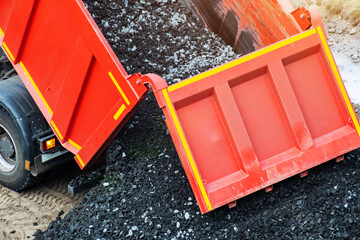 Red dump truck actively unloads crushed stone onto ground surface, bulk material flowing from rear of vehicle, illustrating heavy machinery work on construction site, material delivery. © HENADZY